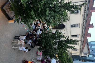 Groupe de personnes rassemblées autour de tables sous des parasols dans une cour extérieure avec végétation.