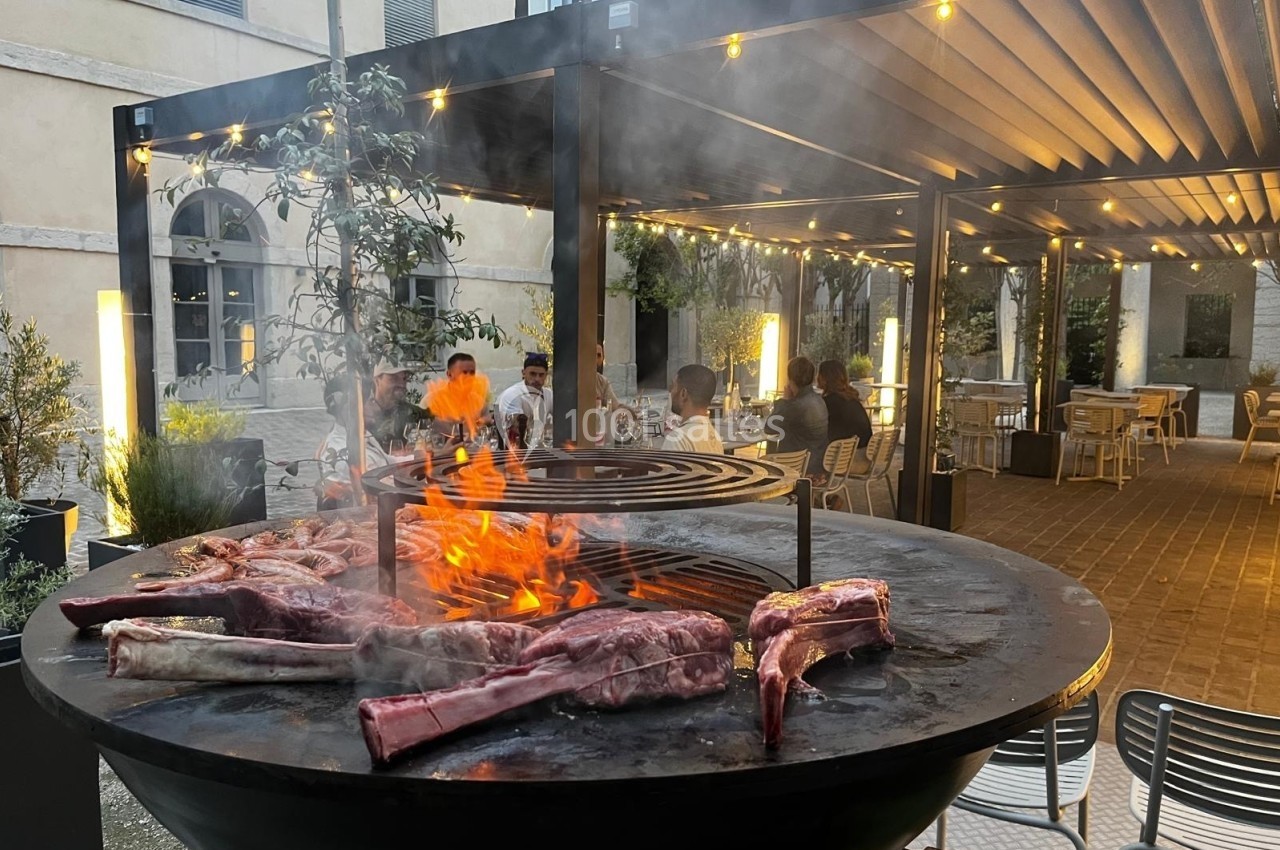 Viande grillant sur un barbecue circulaire en plein air, avec des convives assis sous une pergola éclairée.