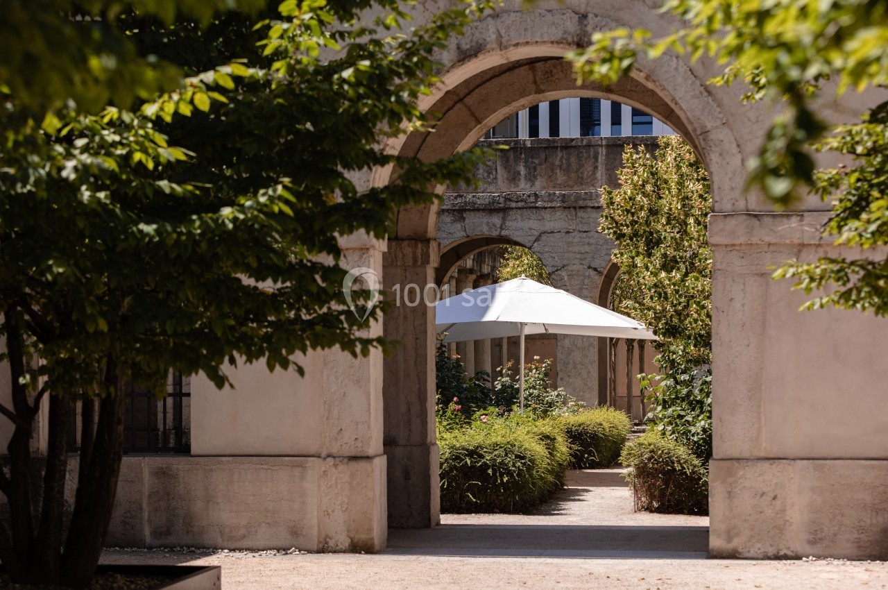 Vue d'une arche en pierre donnant sur un jardin avec des plantes, un chemin et un parasol blanc.