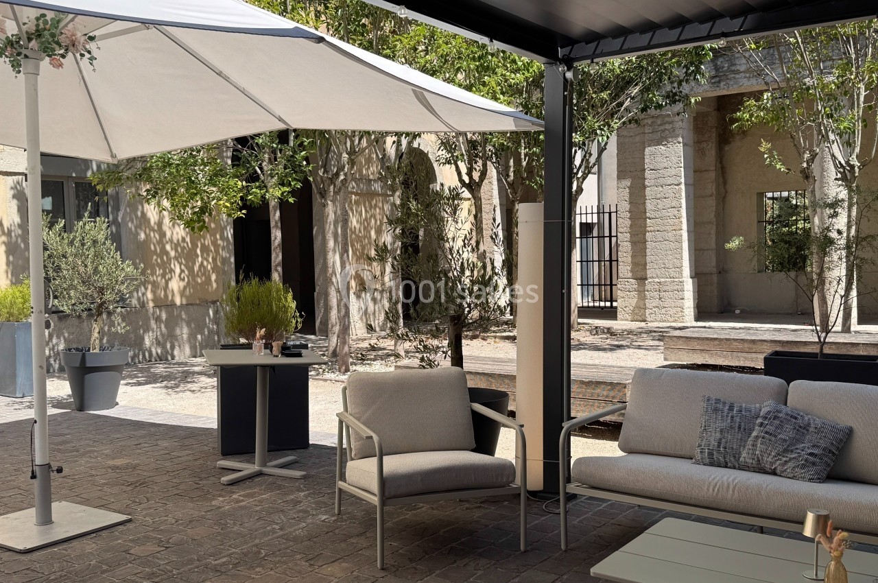 Espace extérieur avec mobilier de jardin sous un parasol, entouré de végétation et de bâtiments en pierre.