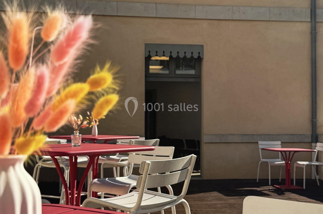 Terrasse avec tables rouges, chaises blanches et vase de fleurs séchées colorées, devant un mur beige avec une porte ouverte.