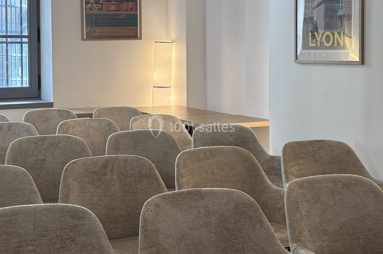 Salle de réunion avec des rangées de chaises beiges, une lampe allumée et des affiches encadrées au mur.