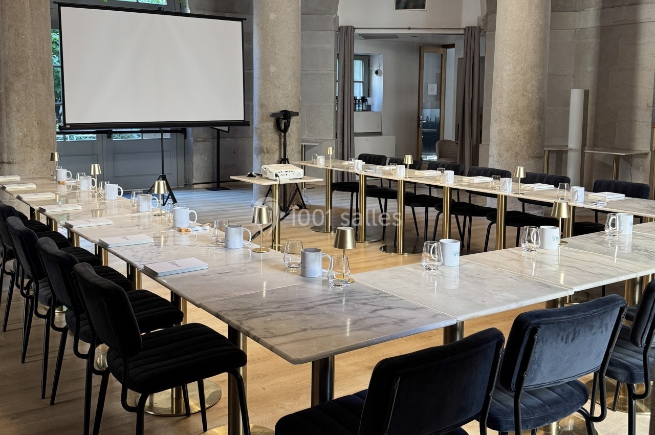 Salle de réunion lumineuse avec des tables en marbre disposées en U, équipée d'un écran de projection et de chaises noires.