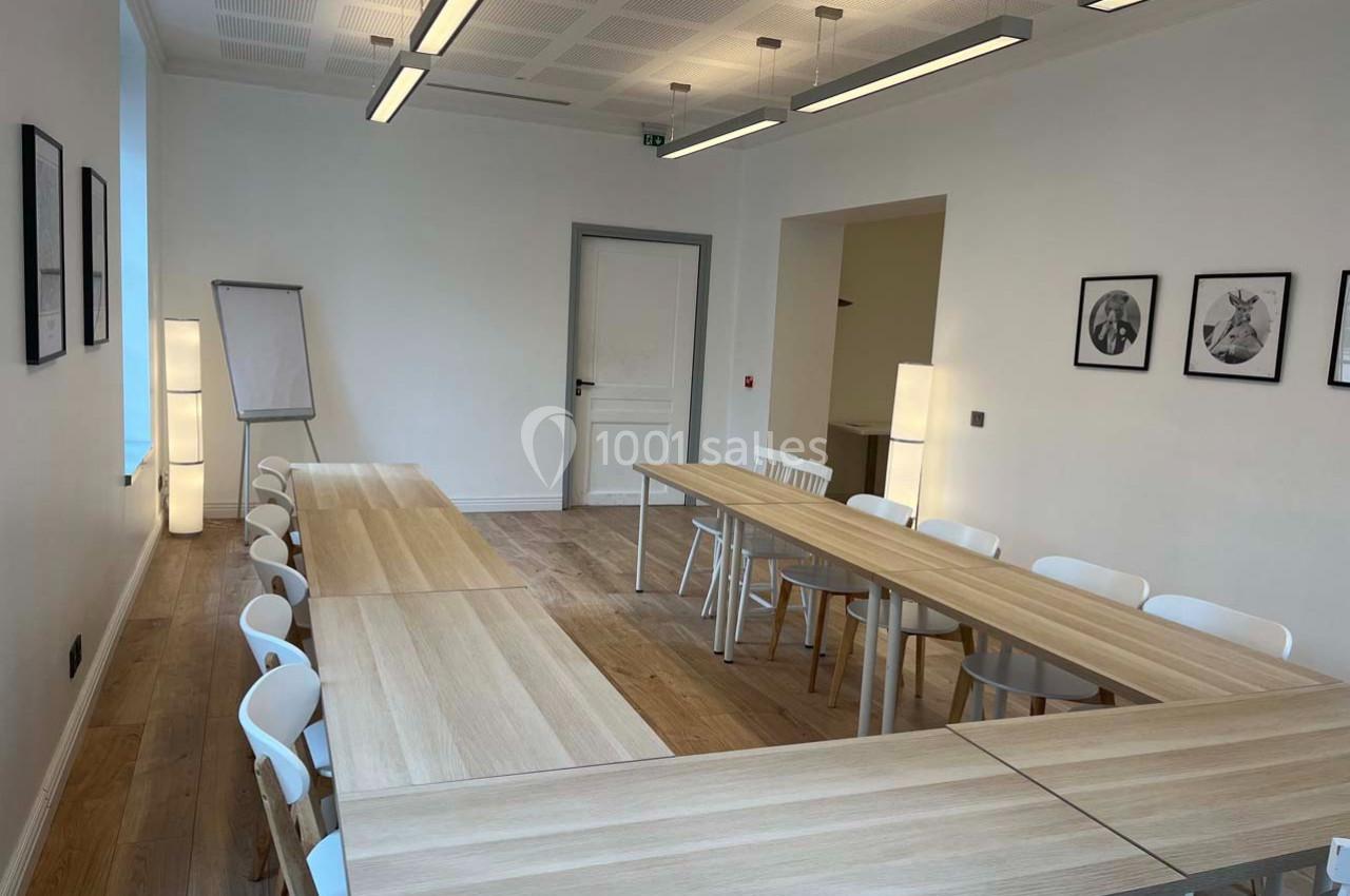Salle de réunion lumineuse avec tables en bois clair disposées en U, chaises blanches et tableaux accrochés au mur.