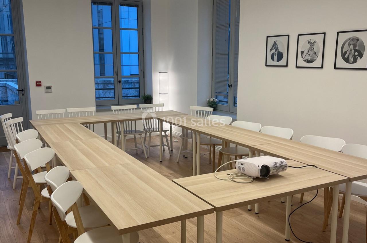 Salle de réunion lumineuse avec tables en bois disposées en U, chaises blanches et projecteur posé sur une table.