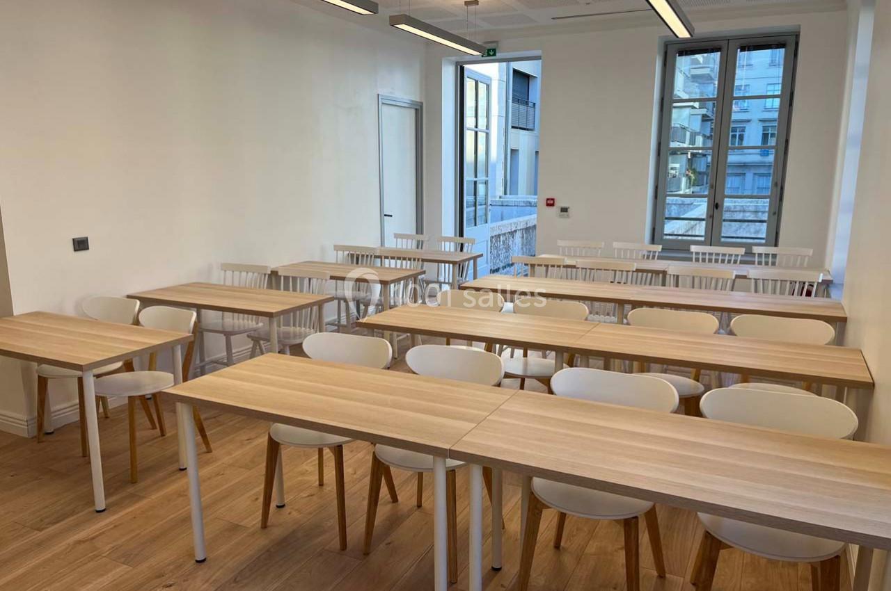 Salle de classe lumineuse avec des tables en bois clair, des chaises blanches et de grandes fenêtres donnant sur l'extérieur.