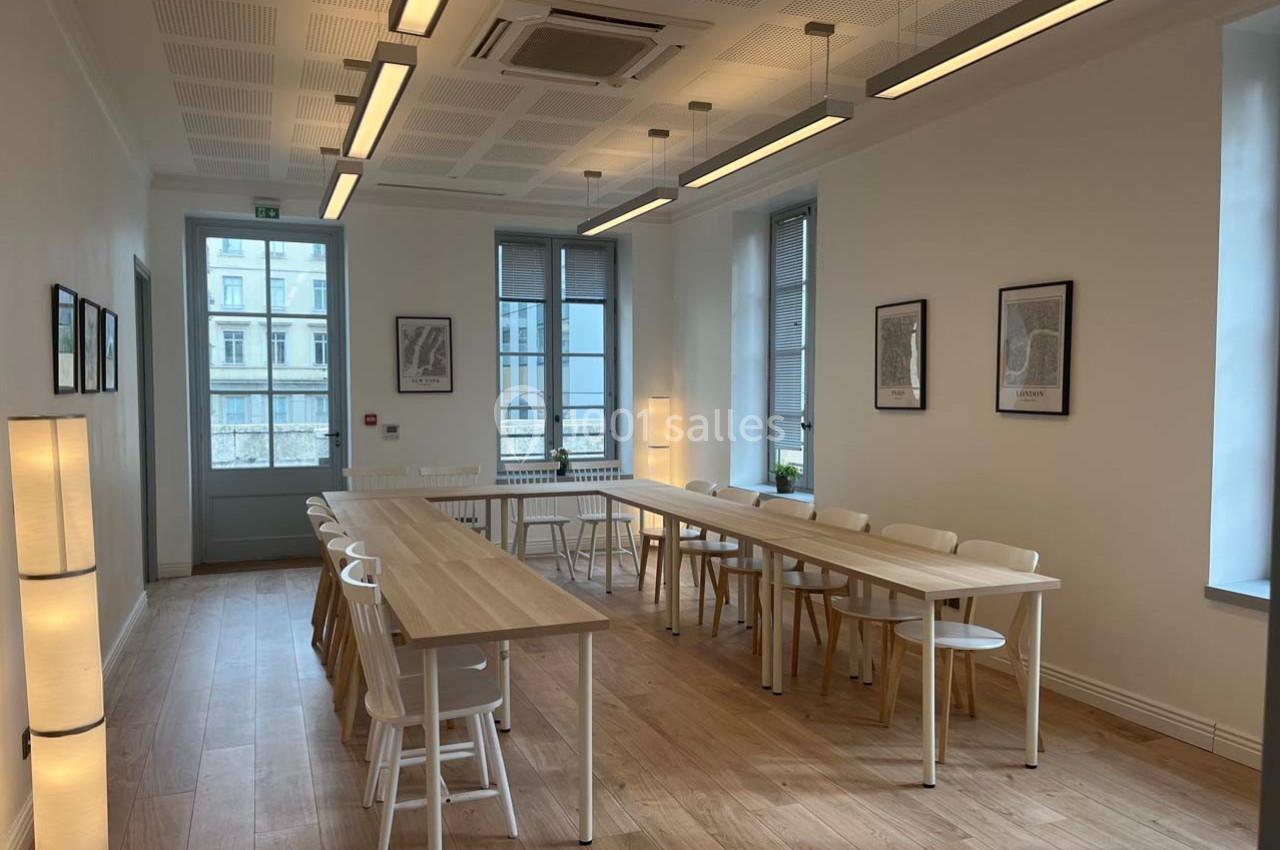 Salle lumineuse avec tables en bois clair, chaises blanches, lampes sur pied et fenêtres donnant sur un bâtiment extérieur.