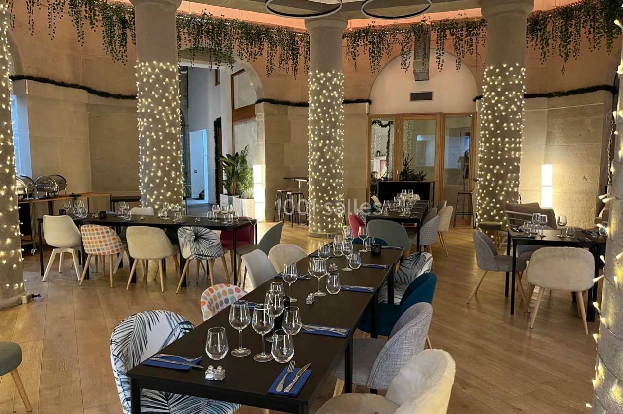 Salle de restaurant élégante avec tables dressées, chaises variées et colonnes décorées de guirlandes lumineuses.