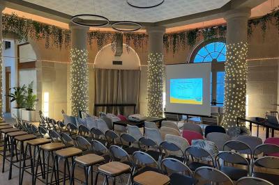 Groupe de personnes rassemblées autour de tables sous des parasols dans une cour extérieure avec végétation.