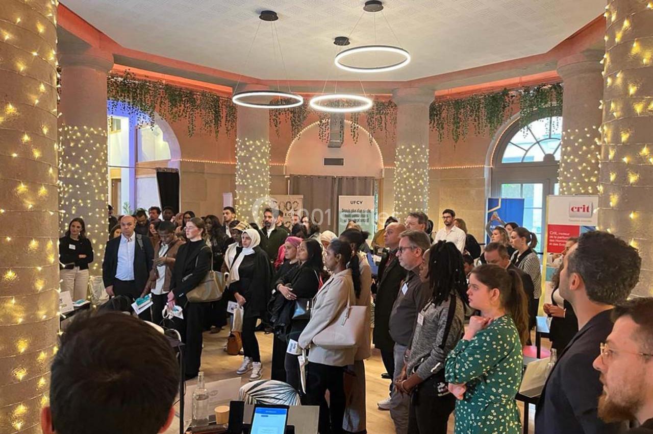 Un groupe de personnes assiste à un événement dans une salle décorée de colonnes lumineuses et de plantes suspendues.