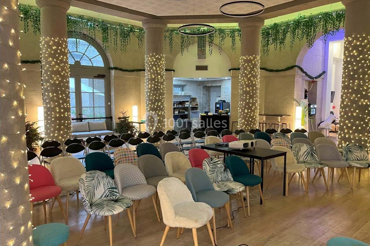 Salle lumineuse avec des rangées de chaises colorées, décorations végétales et guirlandes lumineuses suspendues.