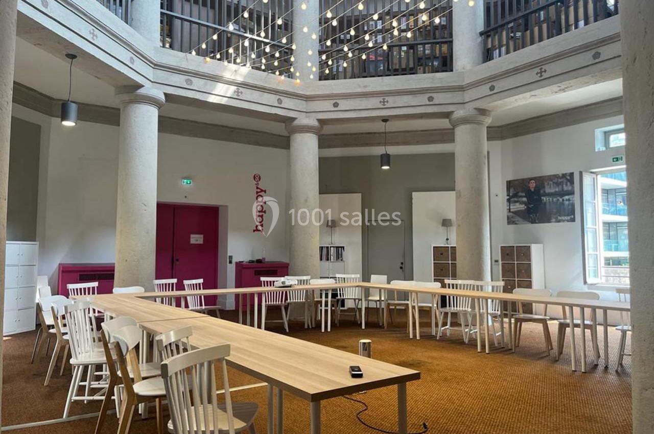 Salle lumineuse avec tables en U, chaises blanches, colonnes, plafond haut et guirlande lumineuse suspendue.