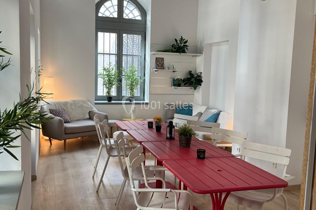 Salle lumineuse avec une grande table rouge entourée de chaises blanches, canapé gris et plantes décoratives près d'une…