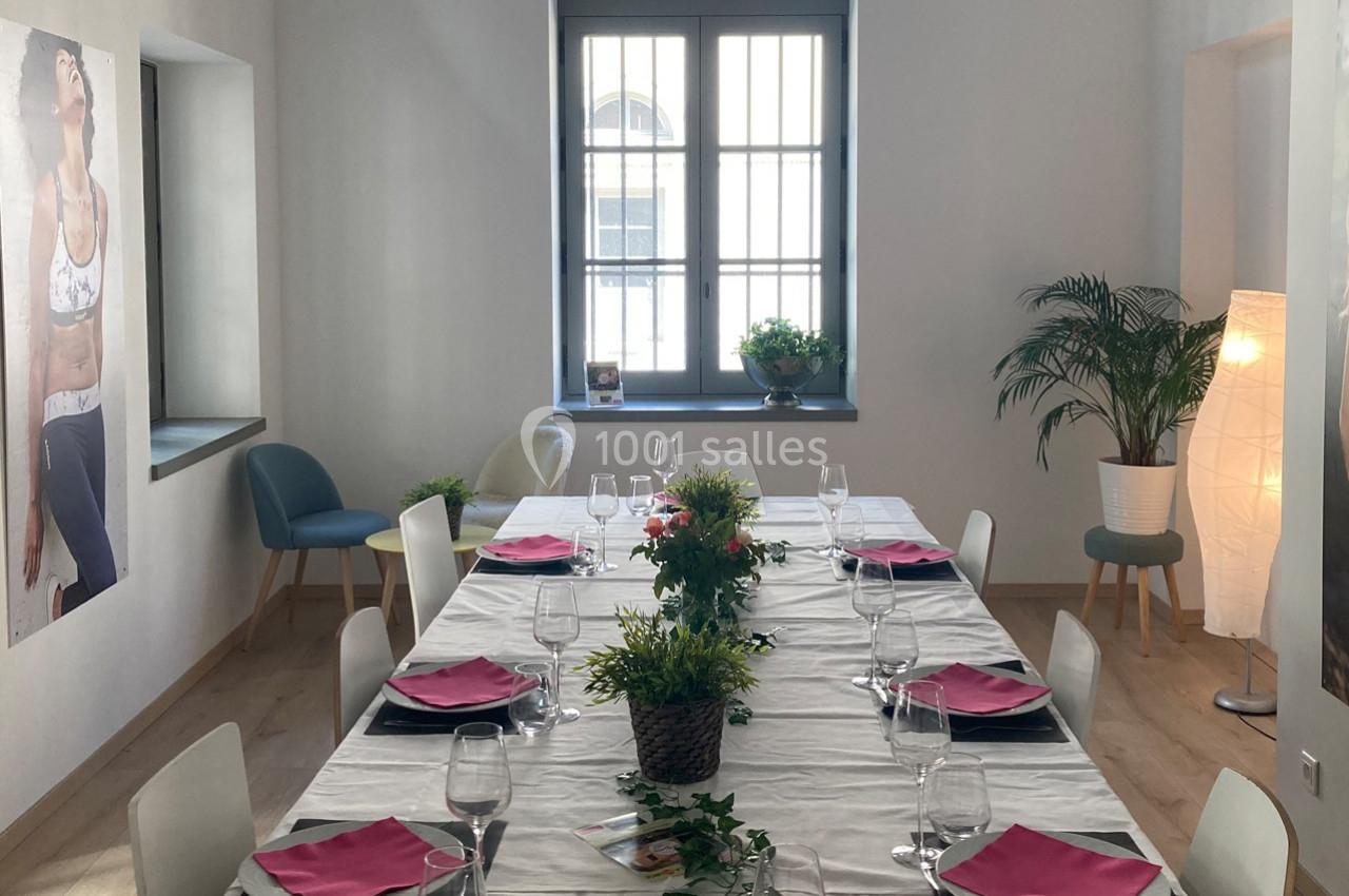 Table dressée avec nappes blanches, serviettes roses, verres et plantes décoratives dans une pièce lumineuse.