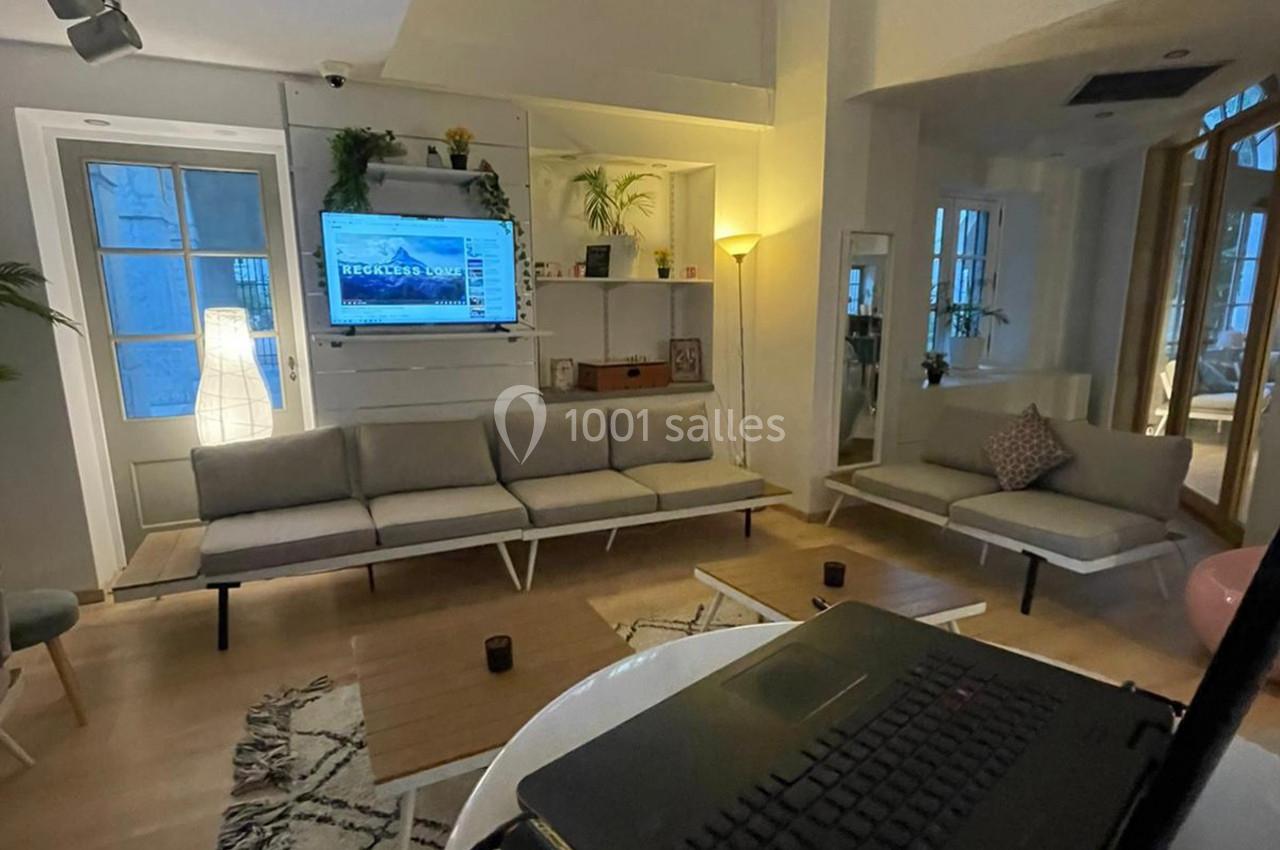 Salon moderne avec canapé beige, table basse en bois, éclairage chaleureux et écran allumé sur un mur blanc.