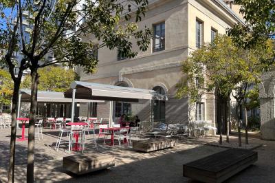 Groupe de personnes rassemblées autour de tables sous des parasols dans une cour extérieure avec végétation.