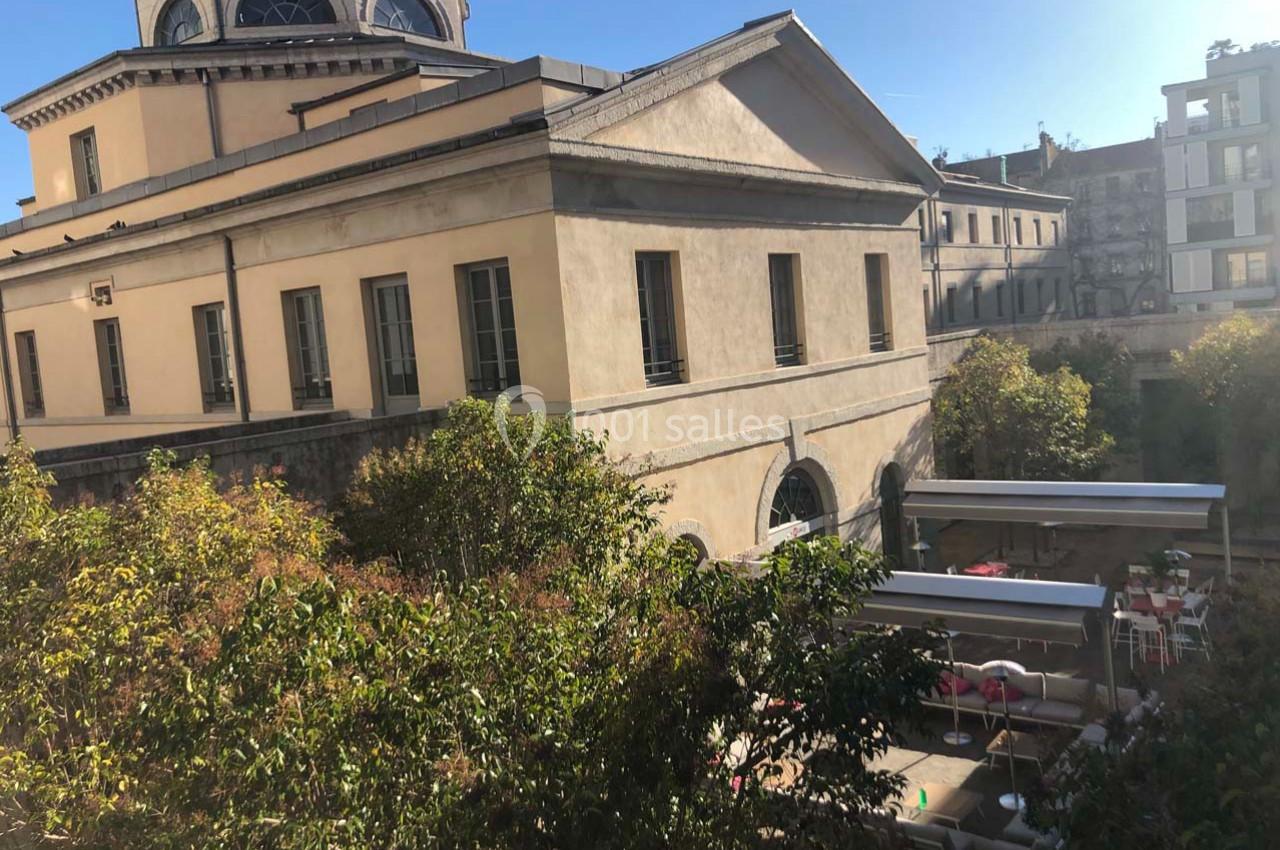 Bâtiment ancien avec façade beige, entouré de végétation et d'une terrasse aménagée avec tables et chaises.
