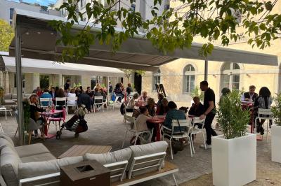 Groupe de personnes rassemblées autour de tables sous des parasols dans une cour extérieure avec végétation.