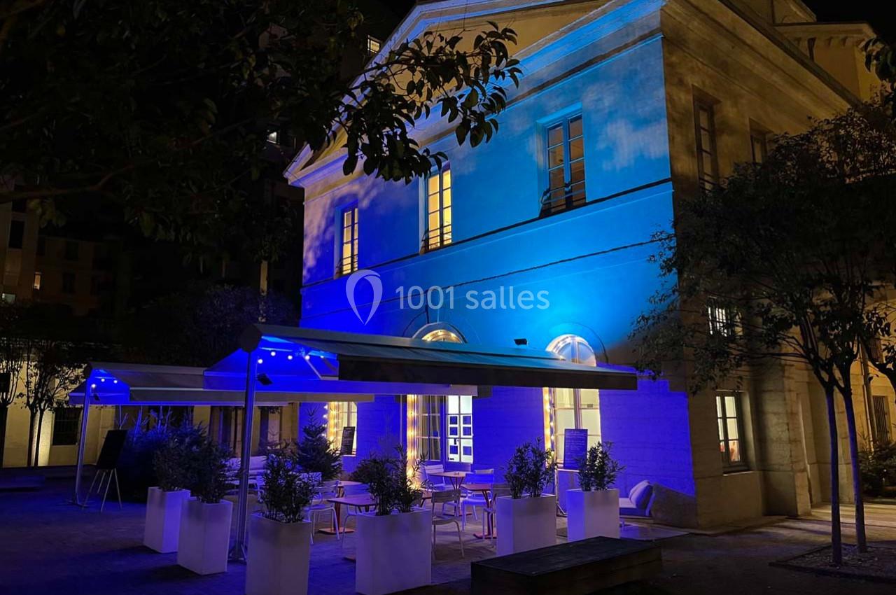 Bâtiment éclairé en bleu la nuit, avec une terrasse aménagée de tables, chaises et plantes en pots.