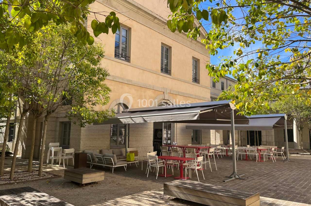 Cour extérieure avec terrasses aménagées, tables et chaises sous des auvents, entourée d'arbres et d'un bâtiment beige.