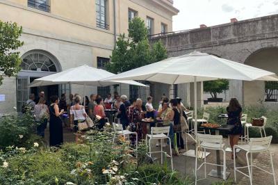 Groupe de personnes rassemblées autour de tables sous des parasols dans une cour extérieure avec végétation.