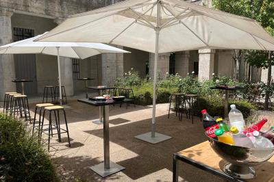 Groupe de personnes rassemblées autour de tables sous des parasols dans une cour extérieure avec végétation.