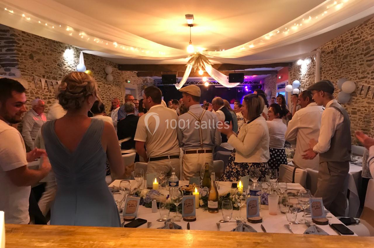 Salle de réception décorée avec des guirlandes lumineuses, des invités debout applaudissant près de tables dressées.