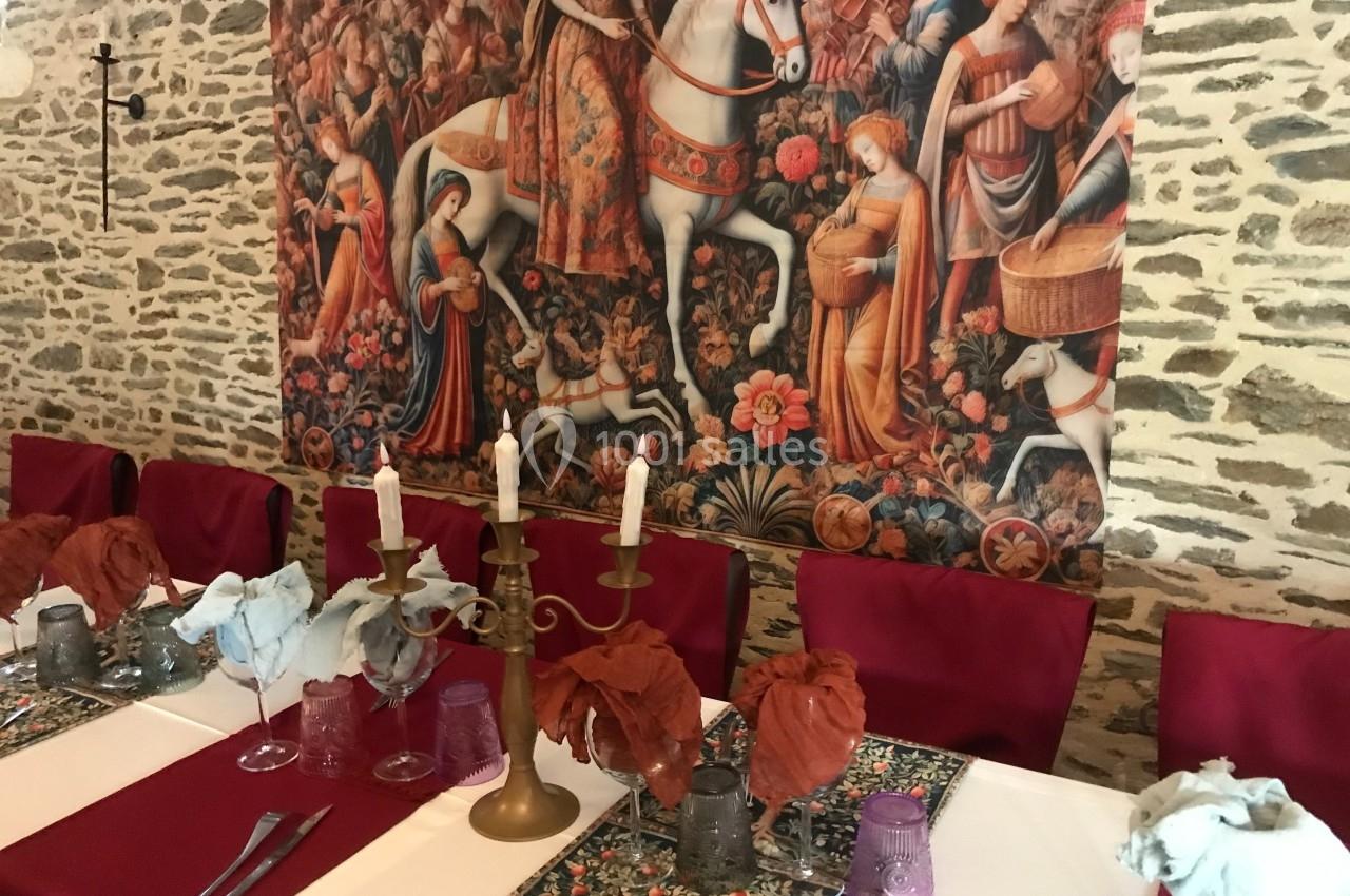 Salle à manger décorée avec une tapisserie médiévale, des chaises rouges et une table dressée avec des chandeliers.