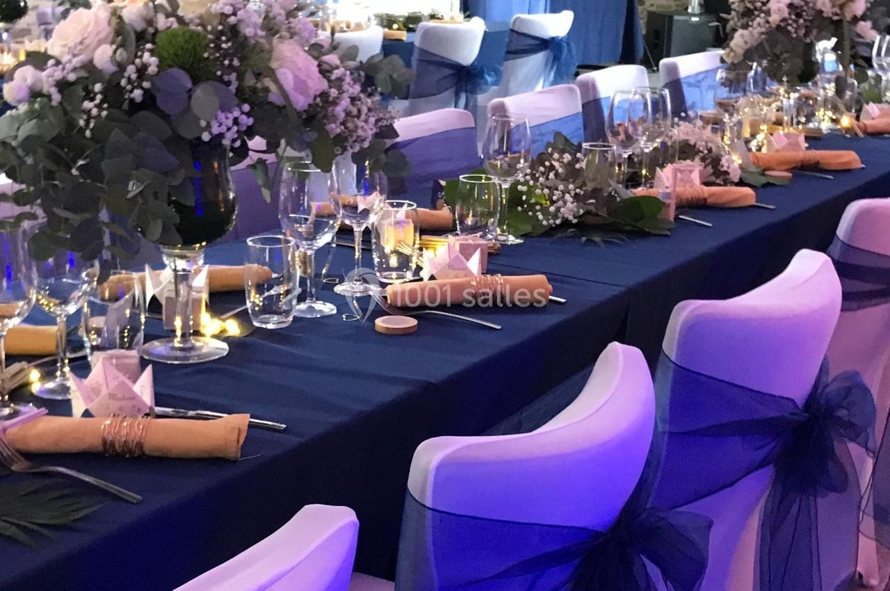 Table de réception élégamment dressée avec nappes bleues, chaises décorées, fleurs blanches et éclairage tamisé.