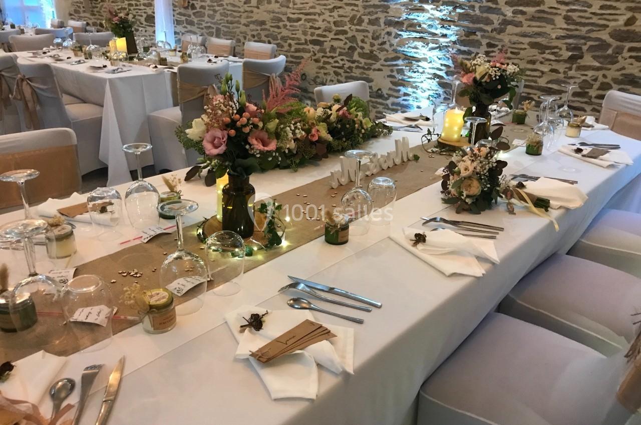 Table de réception décorée avec des fleurs, bougies et nappes blanches, dans une salle aux murs en pierre apparente.