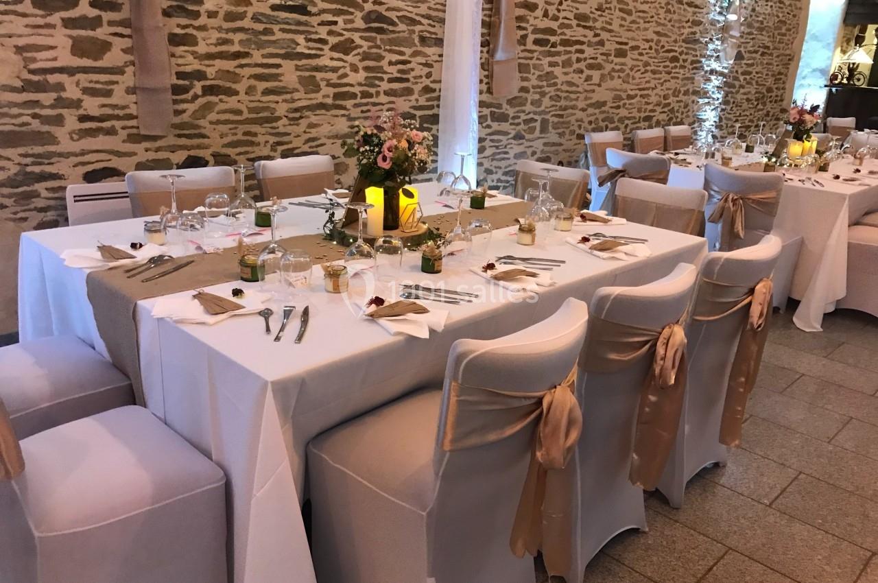 Salle décorée pour un repas, avec tables dressées, nappes blanches, chaises ornées de rubans dorés et murs en pierre…