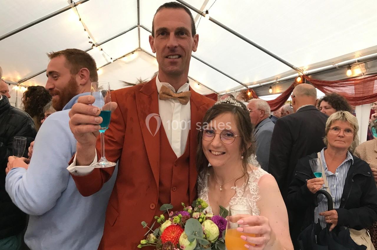 Un couple en tenue de mariage sourit en levant des verres sous une tente éclairée, entouré d'invités.