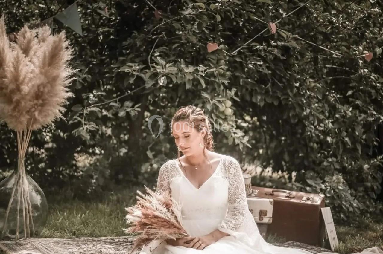 Une femme en robe de mariée est assise sur un tapis dans un jardin, tenant un bouquet de fleurs séchées.