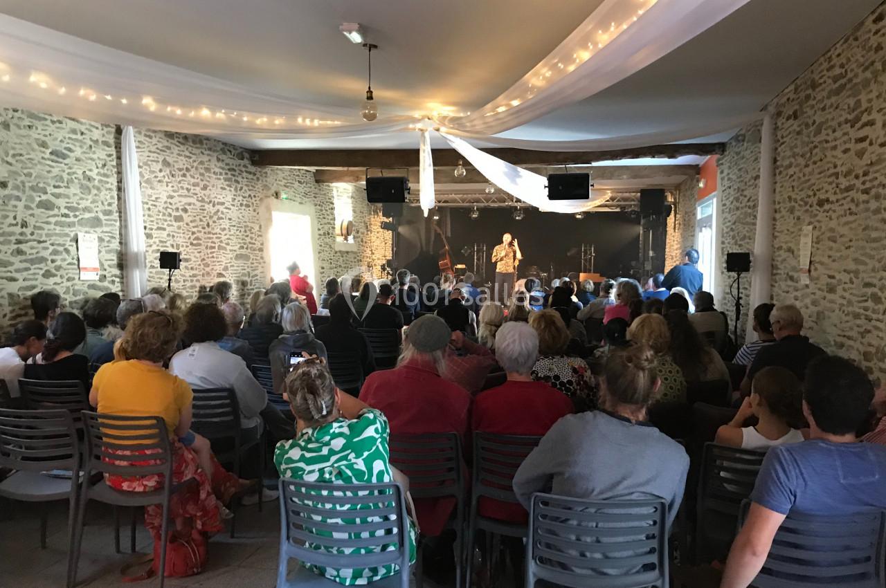 Un groupe de personnes assises dans une salle en pierre, écoutant une performance sur scène sous des guirlandes lumineuses.