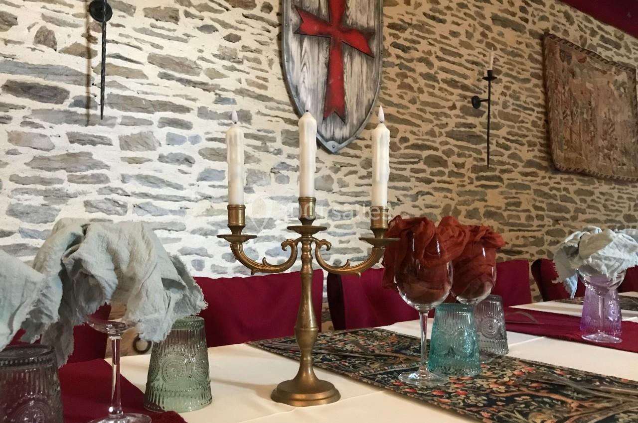 Chandelier en métal doré sur une table dressée, décorée de verres et tissus, dans une salle aux murs en pierre.
