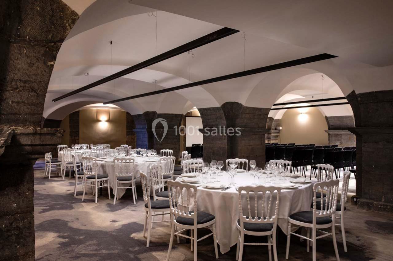Salle voûtée avec tables rondes dressées pour un événement, chaises blanches et éclairage tamisé.