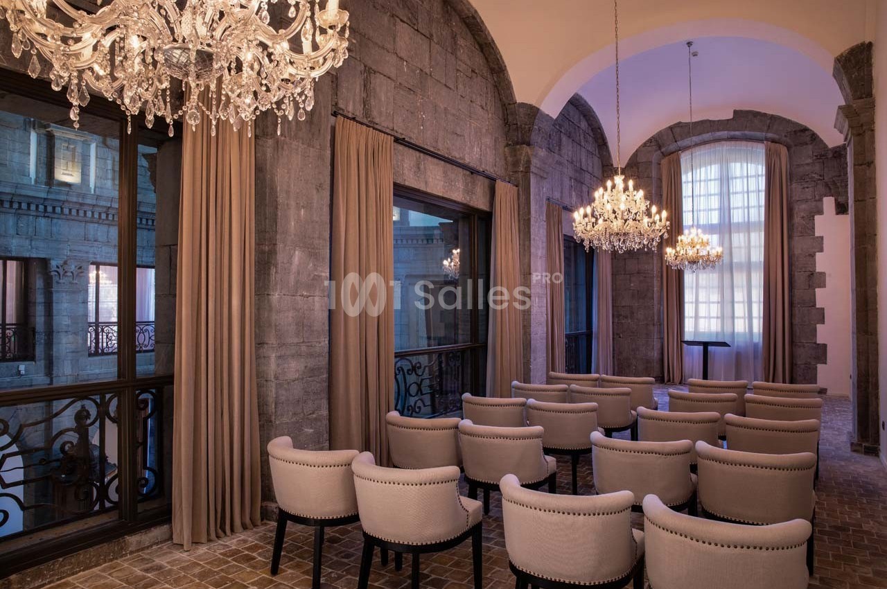 Salle avec des chaises alignées, lustres en cristal suspendus et grandes fenêtres encadrées de rideaux beiges.