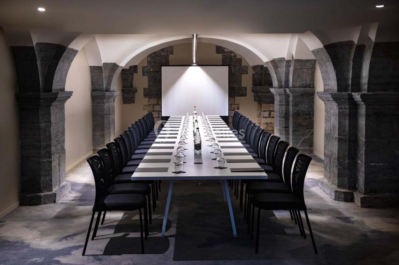 Salle de réunion voûtée avec une longue table rectangulaire, chaises noires alignées et écran de projection en fond.