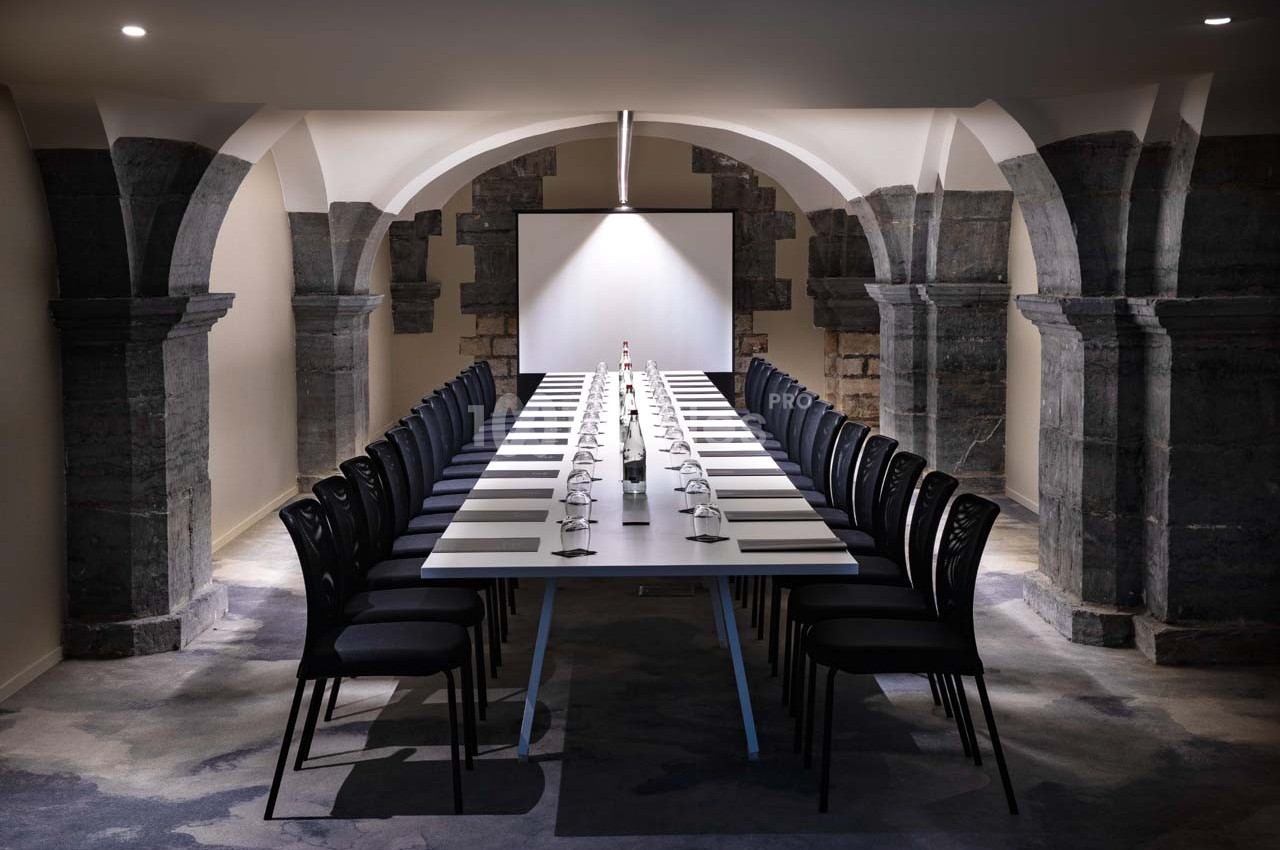 Salle de réunion voûtée avec une longue table rectangulaire, chaises noires alignées et écran de projection en fond.