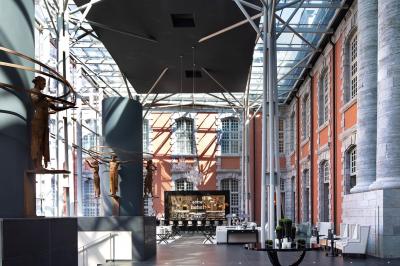 Chambre élégante avec lit double, fauteuil, rideaux beiges, tableaux encadrés et lumière naturelle entrant par une grande…