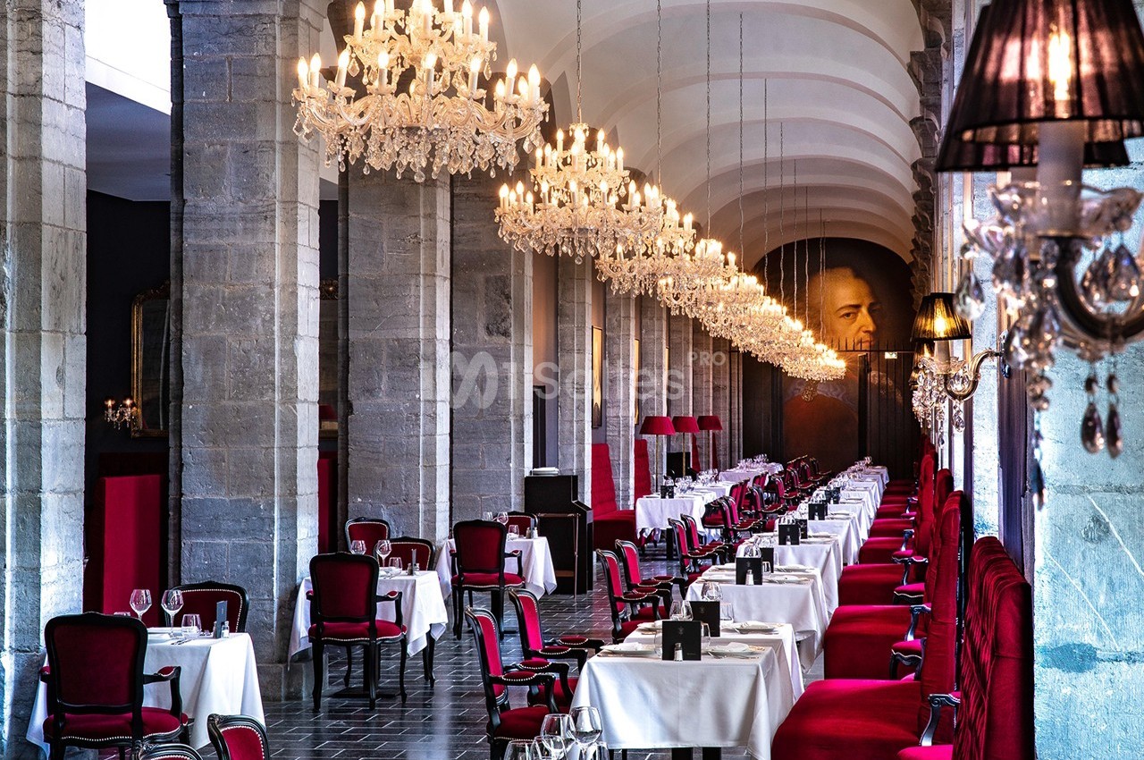 Salle de restaurant élégante avec tables dressées, chaises rouges, lustres en cristal et murs en pierre grise.