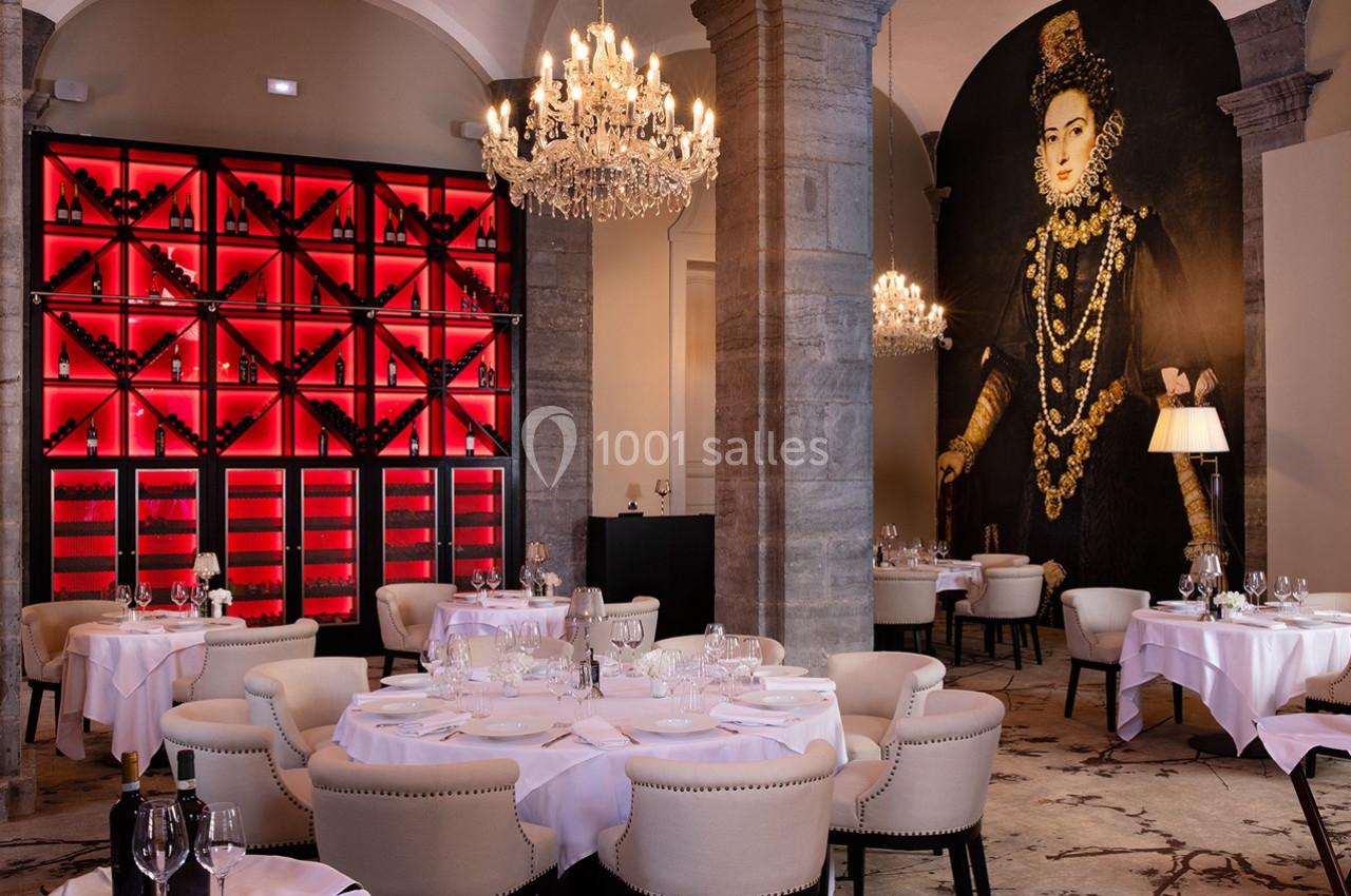 Salle de restaurant élégante avec tables dressées, lustres en cristal, mur rouge à bouteilles et fresque murale ornée.