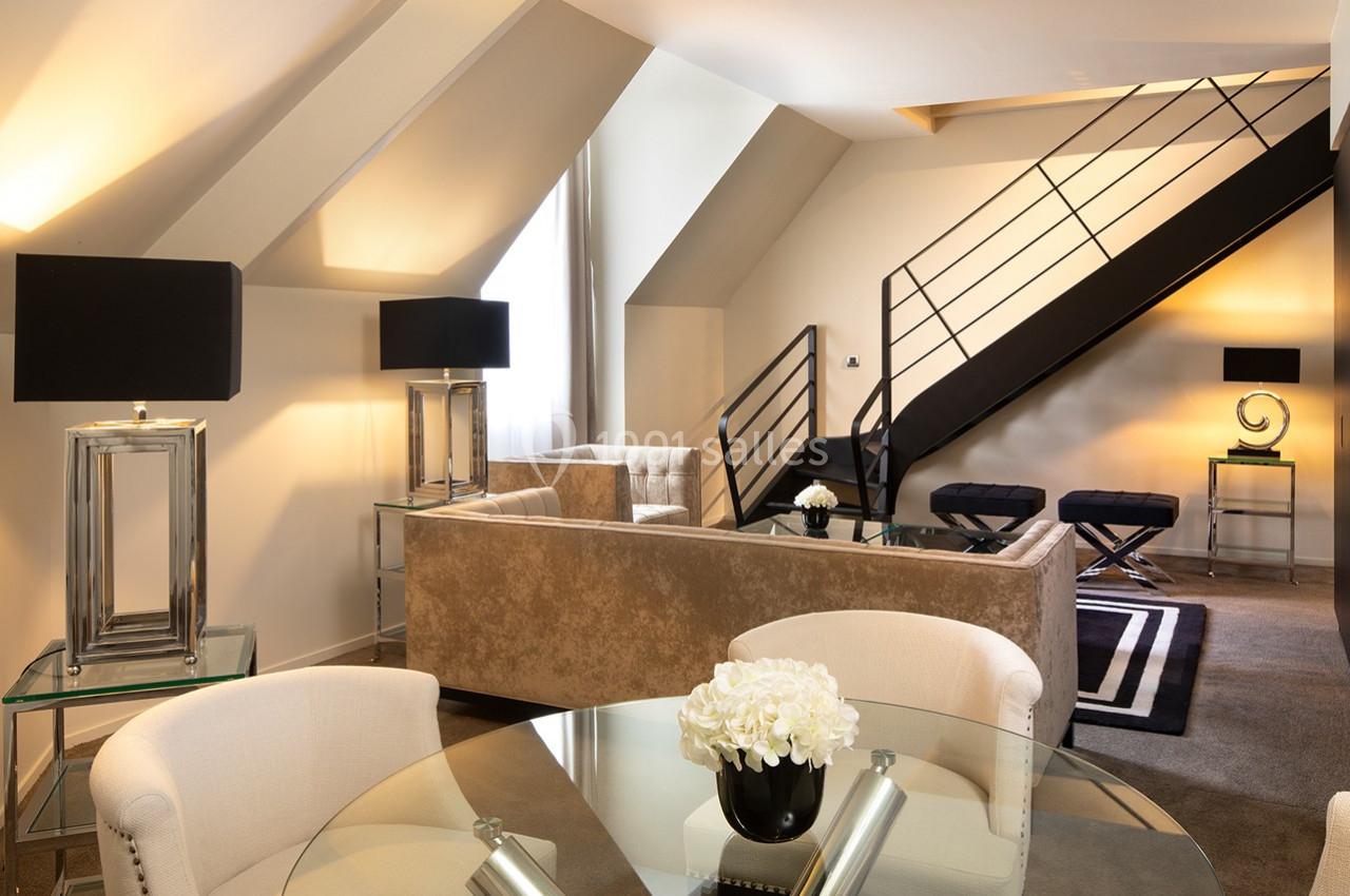 Salon moderne sous combles avec canapé beige, table en verre, lampes noires et escalier menant à une mezzanine.