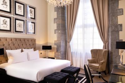 Chambre élégante avec lit double, fauteuil, rideaux beiges, tableaux encadrés et lumière naturelle entrant par une grande…