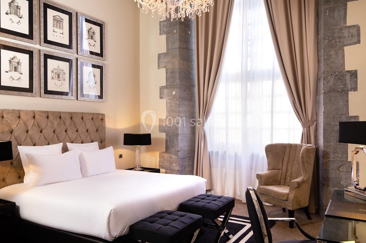 Chambre élégante avec lit double, fauteuil, rideaux beiges, tableaux encadrés et lumière naturelle entrant par une grande…