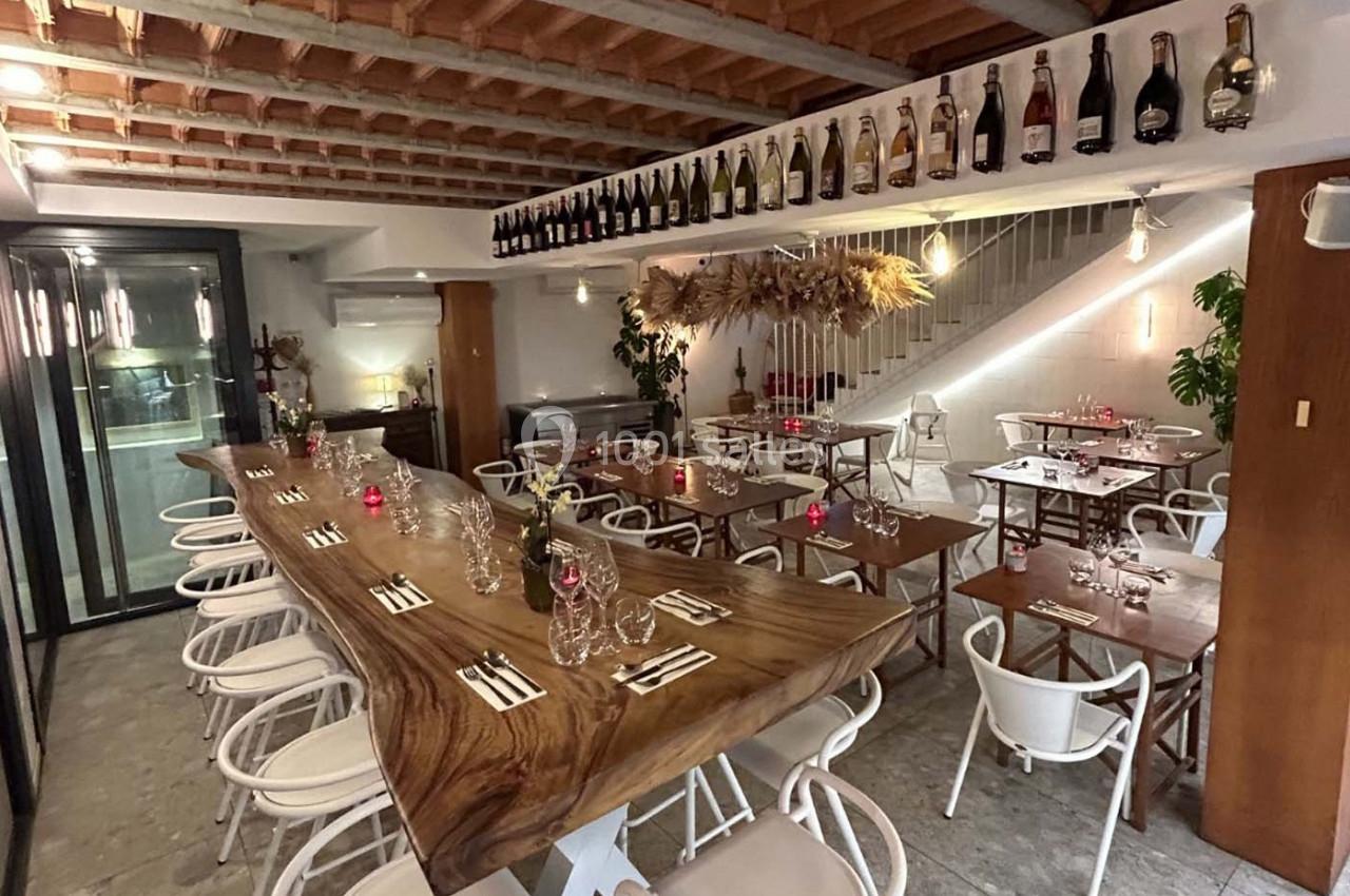 Salle de restaurant moderne avec une grande table en bois, des chaises blanches et des bouteilles exposées en hauteur.