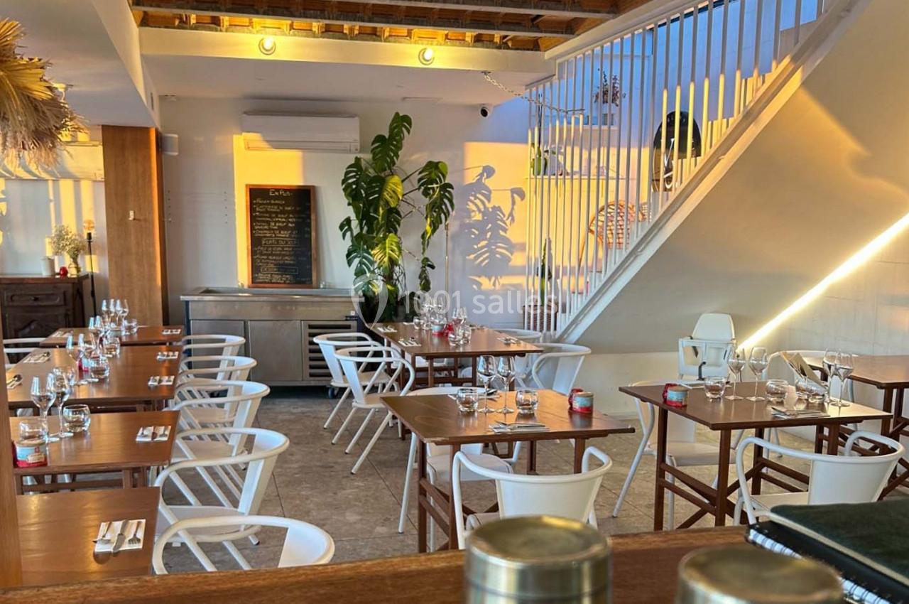 Salle de restaurant lumineuse avec tables en bois, chaises blanches, plantes et tableau noir au mur.