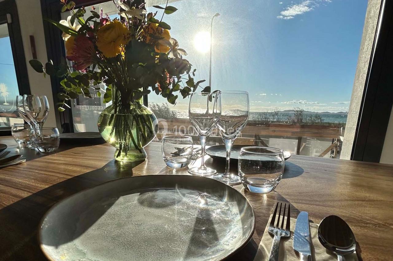 Table dressée avec assiettes, couverts et verres, éclairée par la lumière du soleil près d'une fenêtre avec vue extérieure.