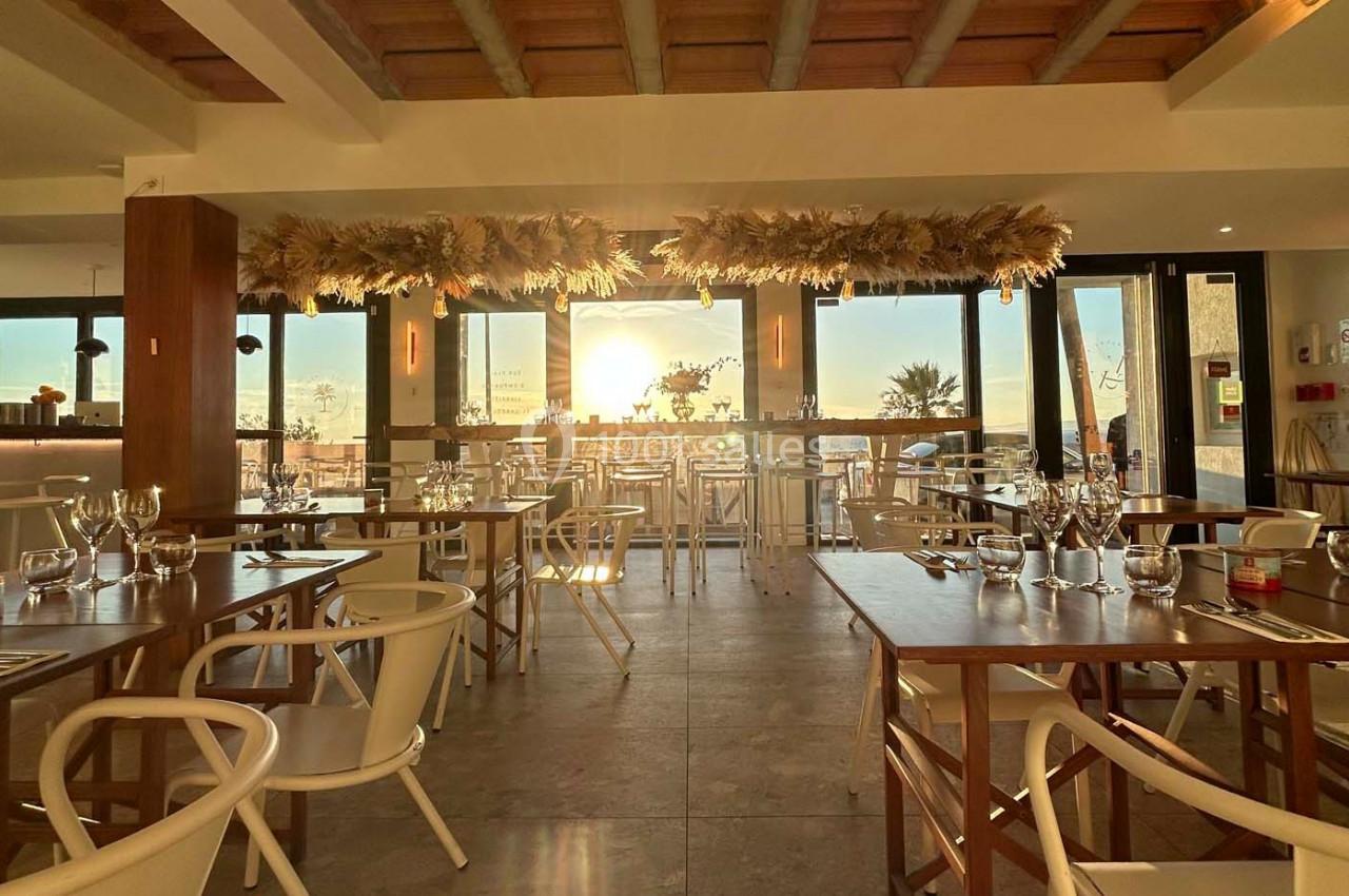 Salle de restaurant lumineuse avec tables en bois, chaises blanches et grande baie vitrée laissant entrer la lumière du…