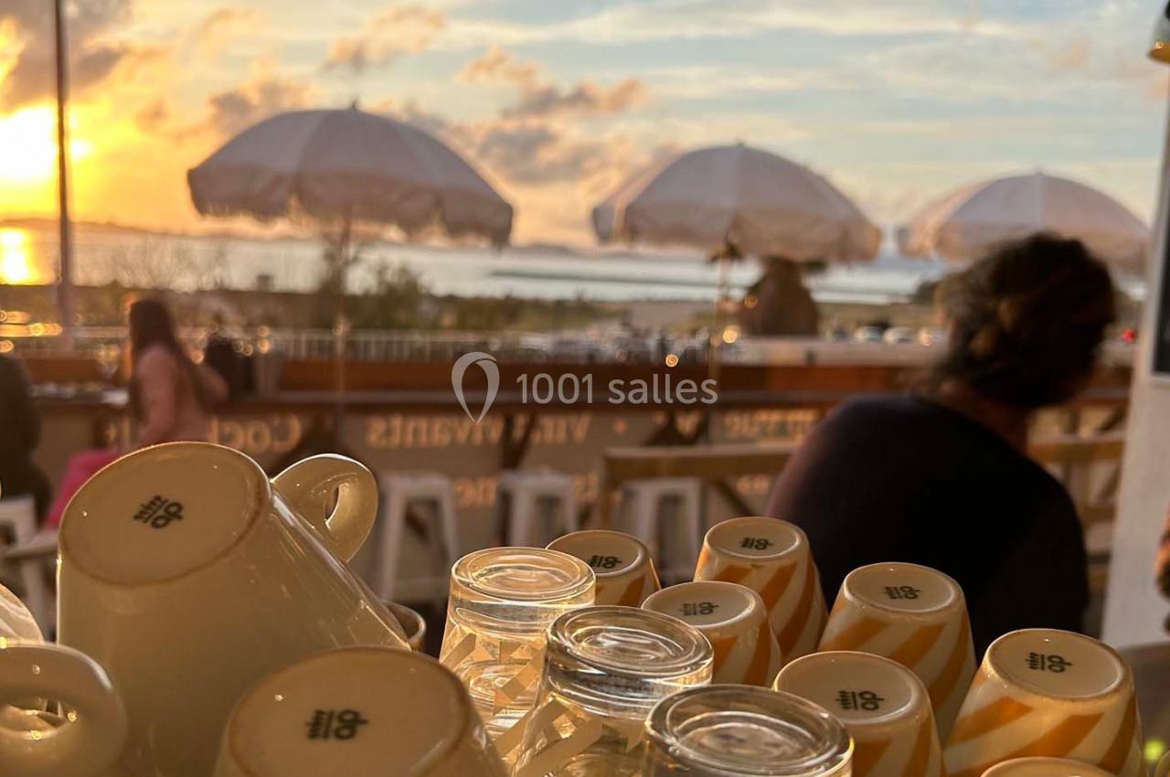 Tasses empilées et verres sur un comptoir en terrasse, avec vue sur un coucher de soleil et des parasols en arrière-plan.